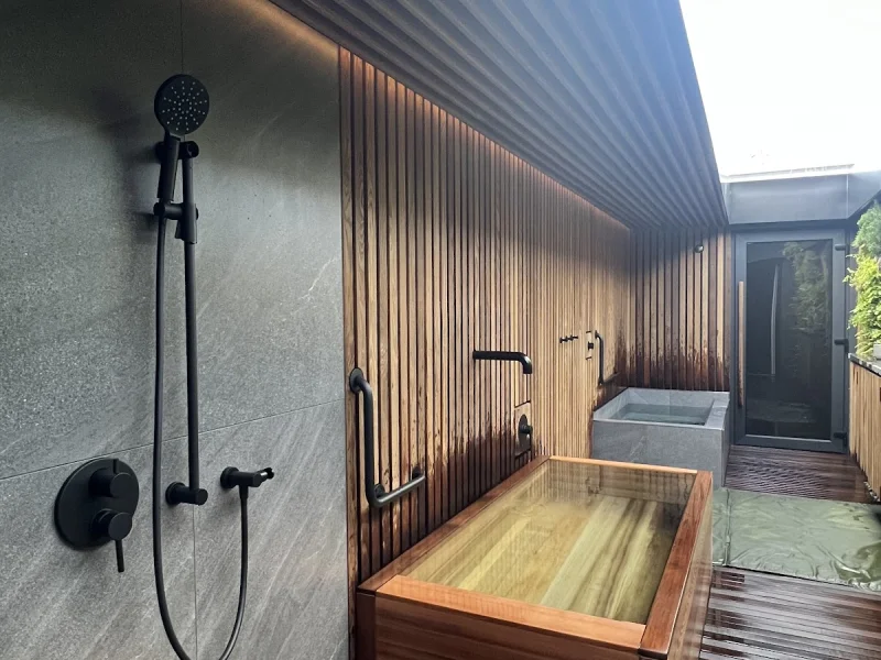 Private outdoor bathing corridor with wooden soaking tub, shower, and dark slatted deck