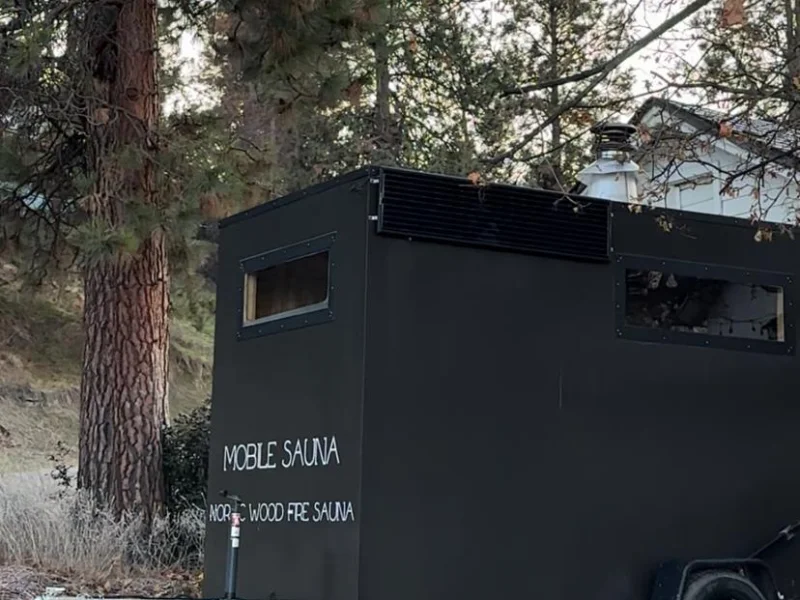 Black mobile sauna trailer parked among tall trees in a forested wellness setting